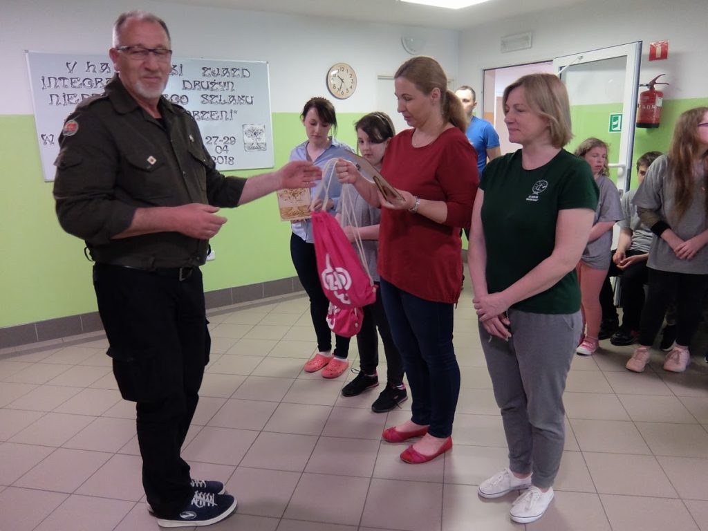 Mężczyzna wręcza nagrodę kobiecie trzymającej różową torbę z prezentami, podczas gdy inni patrzą w jasno oświetlonym pokoju. Grupa ludzi stoi w tle przy ścianie z plakatami i zegarem. Trójka dzieci stoi między nimi, świętując wydarzenie Harcerski Zjazd Integracyjny.